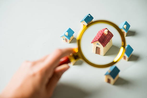 A magnifying glass held by a hand focuses on a miniature model house, symbolizing the process of finding and inspecting affordable home upgrades.