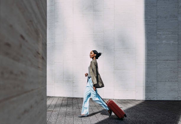 Confident woman walking with a red suitcase in front of a modern urban wall.