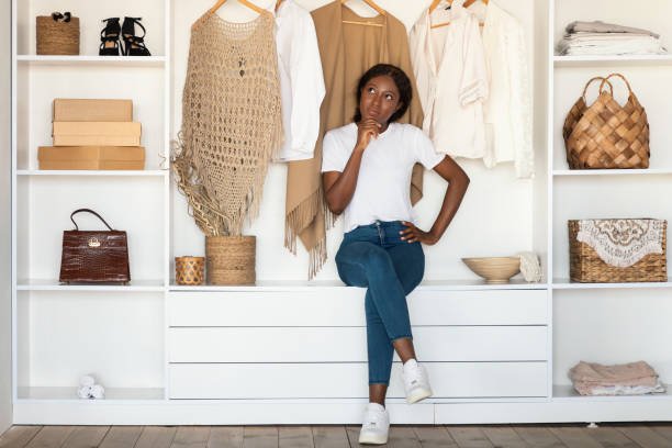 Pensive Black woman sitting among clothes in wardrobe at home.