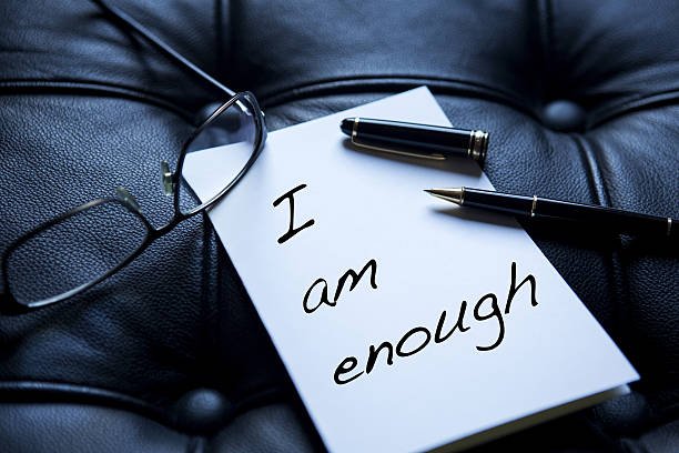 Paper with “I am Enough” written, next to pen and eyeglasses on black leather surface.