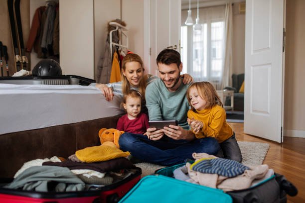 A happy family of four, a mother, father, and two young children, sit together on the floor surrounded by open suitcases, smiling as they look at a tablet to plan their trip