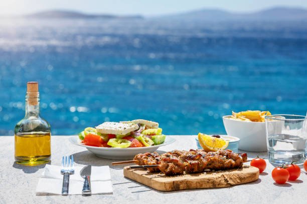 A fresh Greek meal of souvlaki skewers and feta salad on a table overlooking the sparkling blue sea on a sunny day.