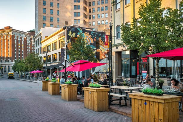  A vibrant city street with people dining at outdoor patios under bright pink umbrellas, illustrating a perfect scene from one of the many fun and affordable Summer Getaways for Less.