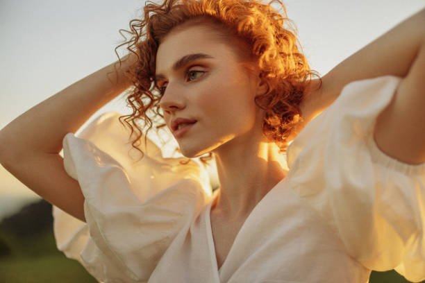 An image illustrating how to live elite on a budget through natural elegance: a close-up portrait of a woman with curly red hair, glowing in warm golden hour sunlight while wearing a simple white blouse.
