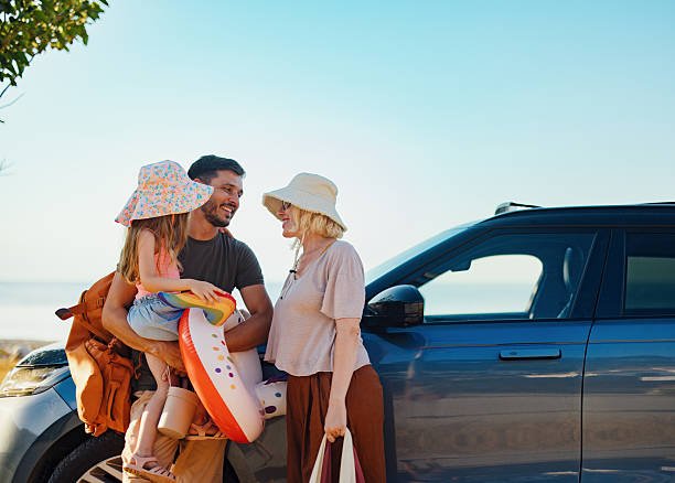 A smiling family—a father holding his young daughter with an inflatable swim ring, and a mother in a sun hat—stand happily next to their SUV on a sunny day, with the water visible in the background, ready for a beach vacation.