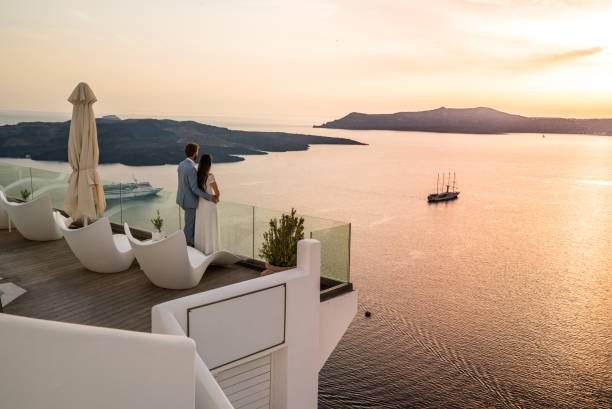 An elegantly dressed couple stands on a modern white balcony, looking out at a stunning sunset over the Santorini caldera. A large cruise ship and a traditional sailing vessel are visible on the calm water below.
