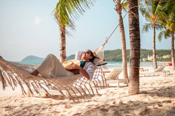 A smiling woman with a straw hat relaxes in a hammock on a tropical beach, enjoying one of the peaceful summer getaways for less.