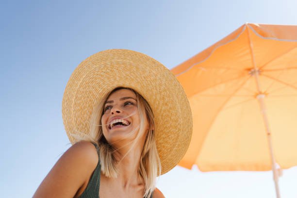 A joyful young woman with blonde hair wearing a large, wide-brimmed straw hat, smiling on a sunny day with a bright orange beach umbrella behind her against a clear blue sky.