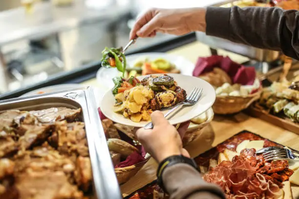 An abundant buffet A person's hands holding a white plate, filling it with various buffet meal items like roasted vegetables and potatoes from a diverse spread.