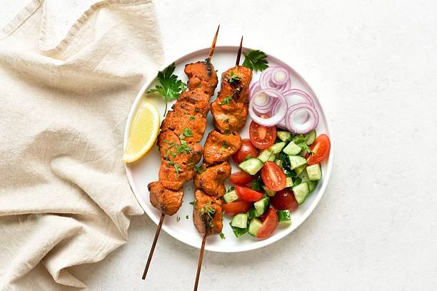 Grilled chicken skewers with a side salad of cucumbers, tomatoes, and red onion rings, garnished with parsley and a lemon wedge, representing delicious summer bbq recipes.