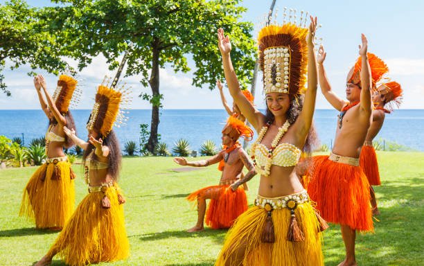 A group of joyful Tahitian dancers in vibrant yellow and orange grass skirts and elaborate shell headdresses performing a traditional dance on a grassy lawn by the sea.