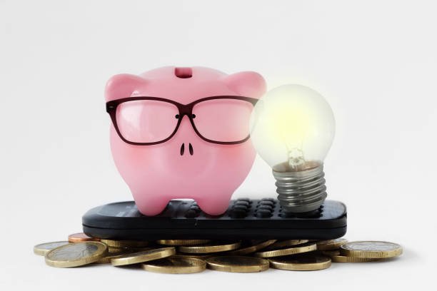 A pink piggy bank wearing glasses sits on a remote control amidst scattered coins, with a lit lightbulb beside it.