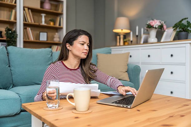A woman sits on the floor of her living room, working on a laptop and reviewing papers at her coffee table.