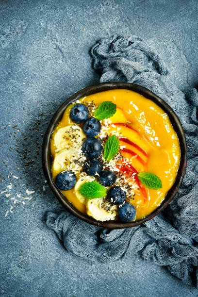 A vibrant tropical mango smoothie bowl in a dark bowl, topped with fresh blueberries, sliced banana, peach, chia seeds, and mint leaves, presented on a blue textured surface.