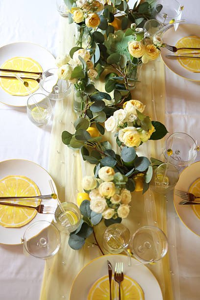 An overhead view of a dining table set for a lemon-themed party. A centerpiece of yellow roses, eucalyptus, and fresh lemons runs down the center, with place settings featuring plates designed to look like lemon slices.