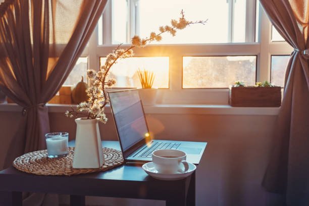 Cozy fall home office décor featuring a laptop and cup of tea on a desk bathed in warm sunset light from a nearby window, creating a serene and productive workspace.