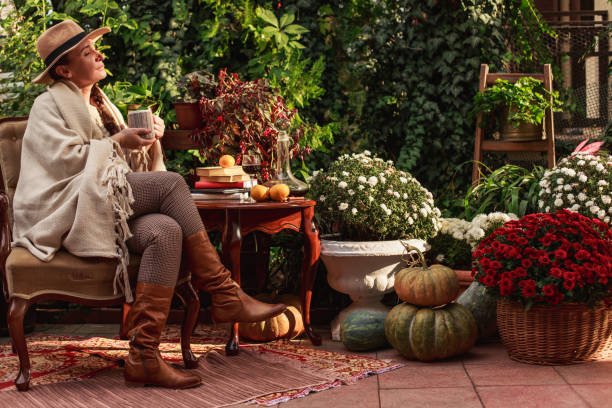 Fall Backyard Oasis2 A serene outdoor patio where a woman has successfully followed tips to create a cozy fall backyard. She is relaxing in a chair wrapped in a warm blanket, surrounded by large pots of colorful mums, pumpkins, and a patterned rug, enjoying a perfect autumn retreat.