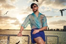 Steal Bad Bunny's Style" - Bad Bunny poses confidently on a boat at sunset, wearing a translucent blue-green button-up shirt with bird motifs, blue shorts, and dark sunglasses. Several seagulls fly around him, and a city skyline is visible in the distance across the water.