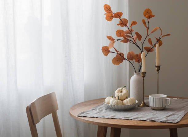 A cozy fall home decor scene featuring a small wooden round table with a light grey linen runner. On the runner, there's a white ceramic bowl filled with small white decorative pumpkins, two tall brass candlesticks with cream-colored taper candles, a white striped mug, and a tall white vase holding rust-colored artificial autumn foliage. A light wooden chair is pulled up to the table, and sheer white curtains hang in the background, letting in soft natural light, creating an inviting atmosphere."