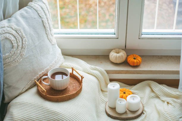 A cozy fall home decor scene featuring a small wooden round table with a light grey linen runner. On the runner, there's a white ceramic bowl filled with small white decorative pumpkins, two tall brass candlesticks with cream-colored taper candles, a white striped mug, and a tall white vase holding rust-colored artificial autumn foliage. A light wooden chair is pulled up to the table, and sheer white curtains hang in the background, letting in soft natural light, creating an inviting atmosphere."