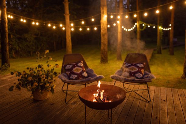 outdoor A cozy Outdoor Fall Décor scene on a wooden deck at night, featuring a glowing fire pit between two chairs with patterned pillows. Strings of warm lights hang in the wooded background, creating a magical and inviting ambiance.