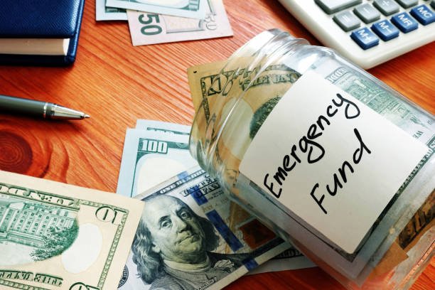 Desk with cash, calculator, and a glass jar labeled "Emergency Fund" overflowing with US dollar bills.