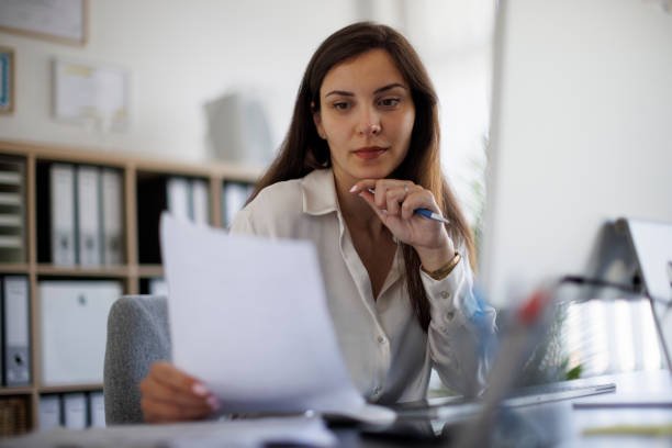 A focused, medium close-up shot of a woman with long dark hair sitting at a desk in a well-organized office. She is wearing a light-colored blouse and intently studying a paper document she holds in her left hand, with her right hand resting near her chin, suggesting concentration or financial planning. A computer monitor is visible in the background, along with organized binders on shelves. The image captures the feeling of analyzing finances or bills, often associated with the phrase "paycheck to paycheck