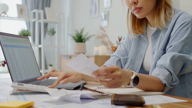  A focused, medium close-up shot of a woman with long dark hair sitting at a desk in a well-organized office. She is wearing a light-colored blouse and intently studying a paper document she holds in her left hand, with her right hand resting near her chin, suggesting concentration or financial planning. A computer monitor is visible in the background, along with organized binders on shelves. The image captures the feeling of analyzing finances or bills, often associated with the phrase "paycheck to paycheck