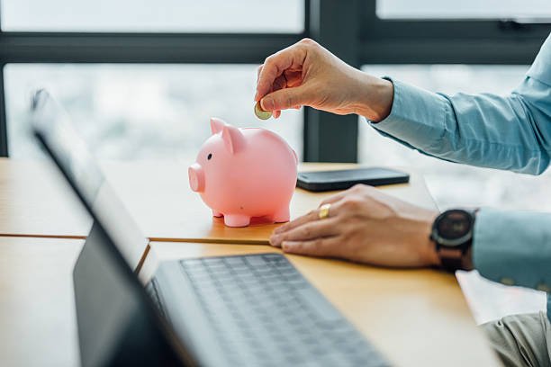 Man putting a coin into a pink piggy bank on a desk next to a laptop, symbolizing saving and starting side income ideas.